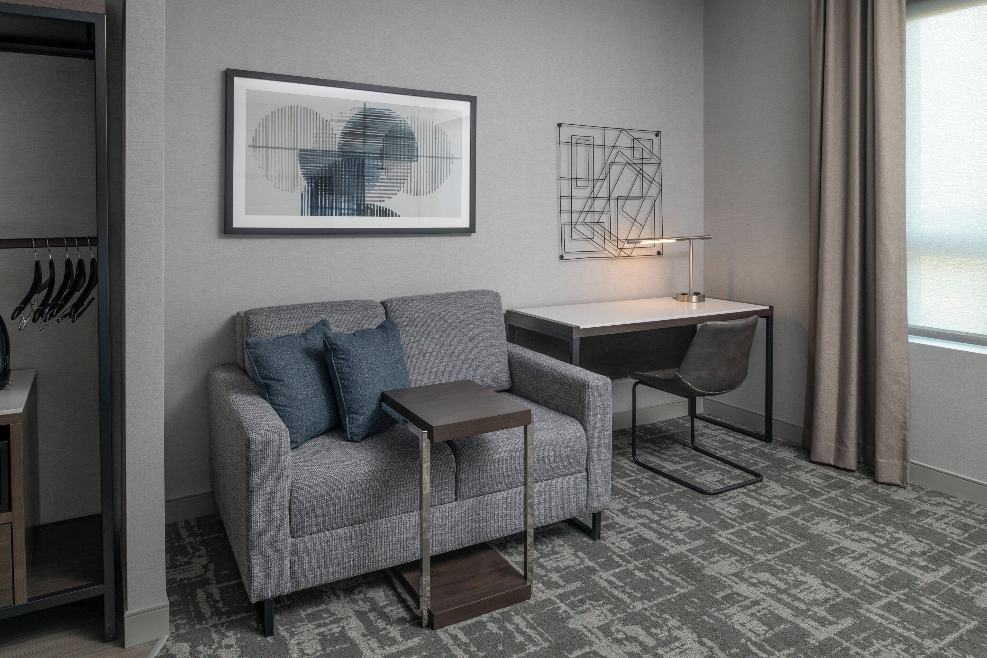 A modern hotel room with a grey sofa, a small side table, a desk with a chair, and a wall-mounted light fixture.