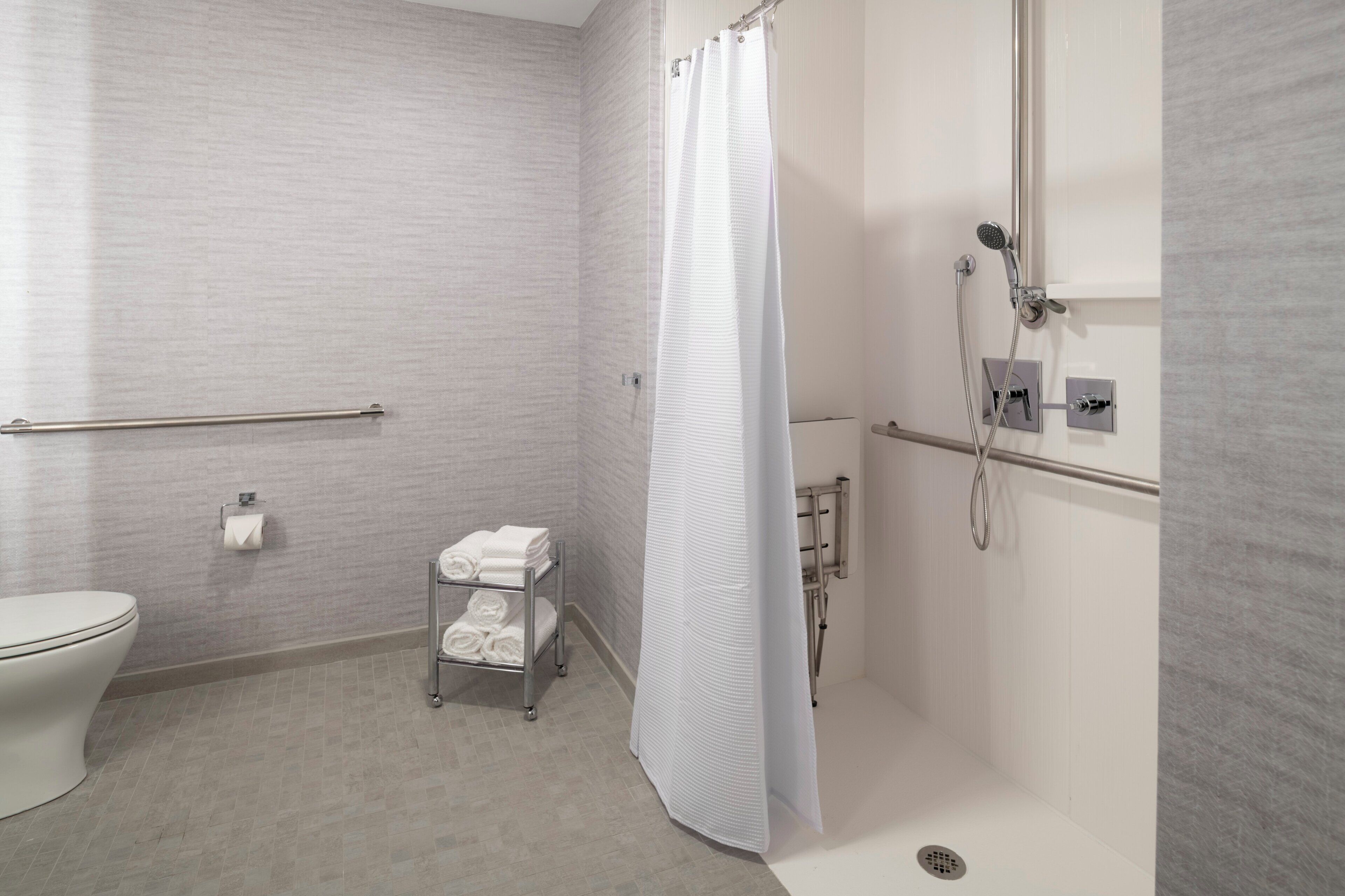 Een badkamer met een toilet, een handdoekrek met opgevouwen handdoeken, een douche met gordijn en een handgreep.