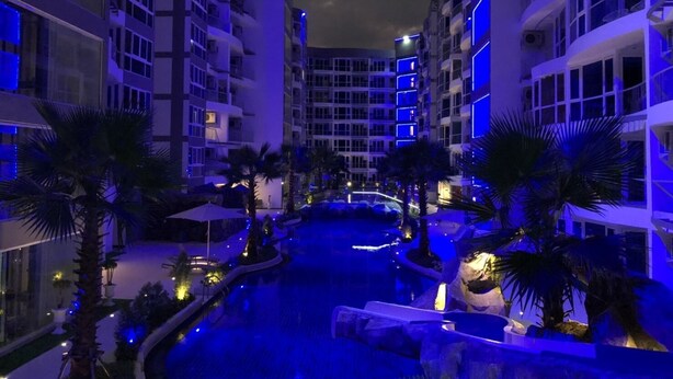 Un hotel moderno con una piscina central, rodeado de palmeras e iluminado con luces azules.
