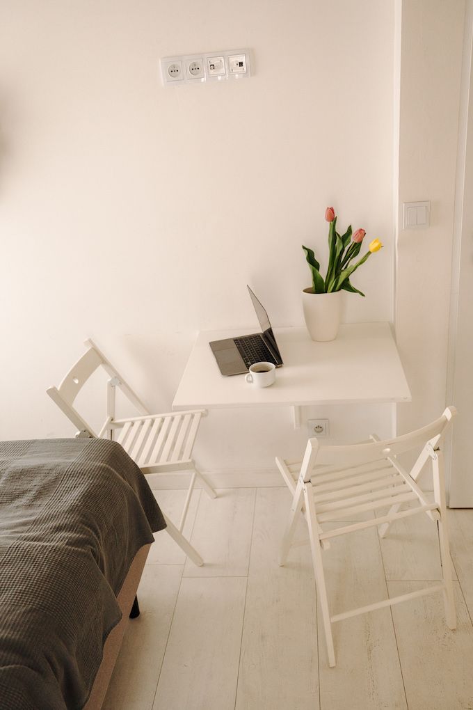 Ein schlicht eingerichteter Raum mit einem Bett, einem Schreibtisch mit Laptop, einer Topfpflanze und zwei weißen Stühlen.
