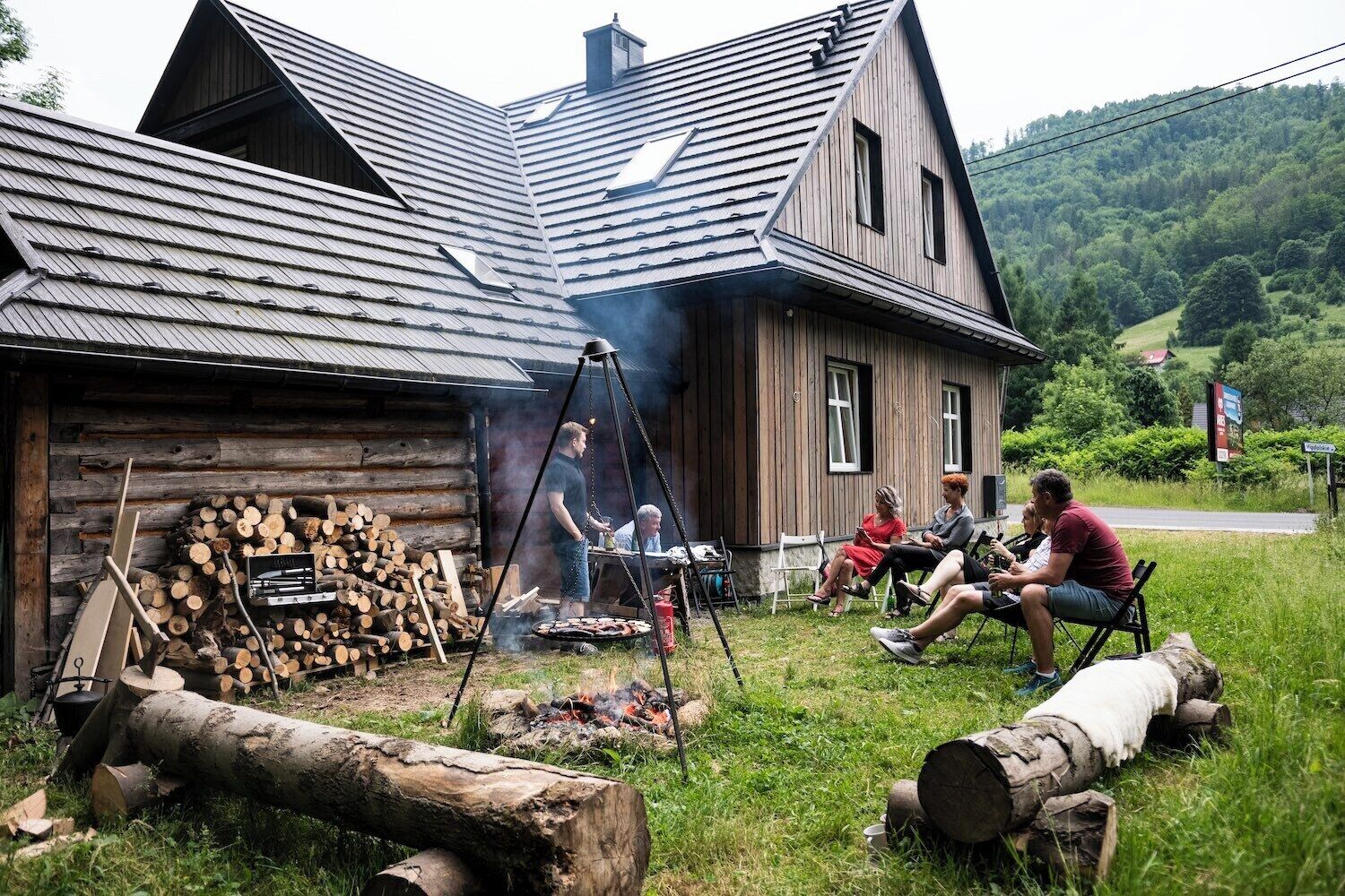 A rustic cabin with a metal roof, a woodpile, and a grill area where people are gathered.
