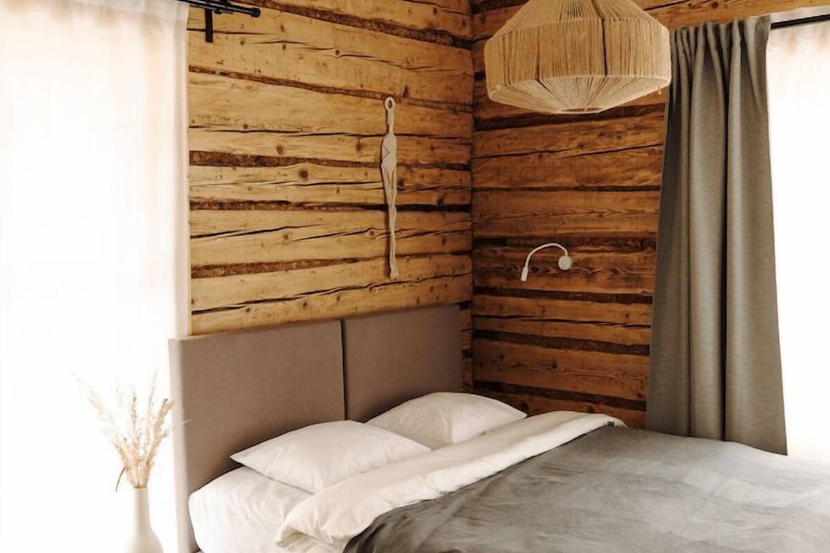 A bedroom with a wooden log wall, a bed with a grey blanket, a hanging light fixture, and a bedside table with a vase.