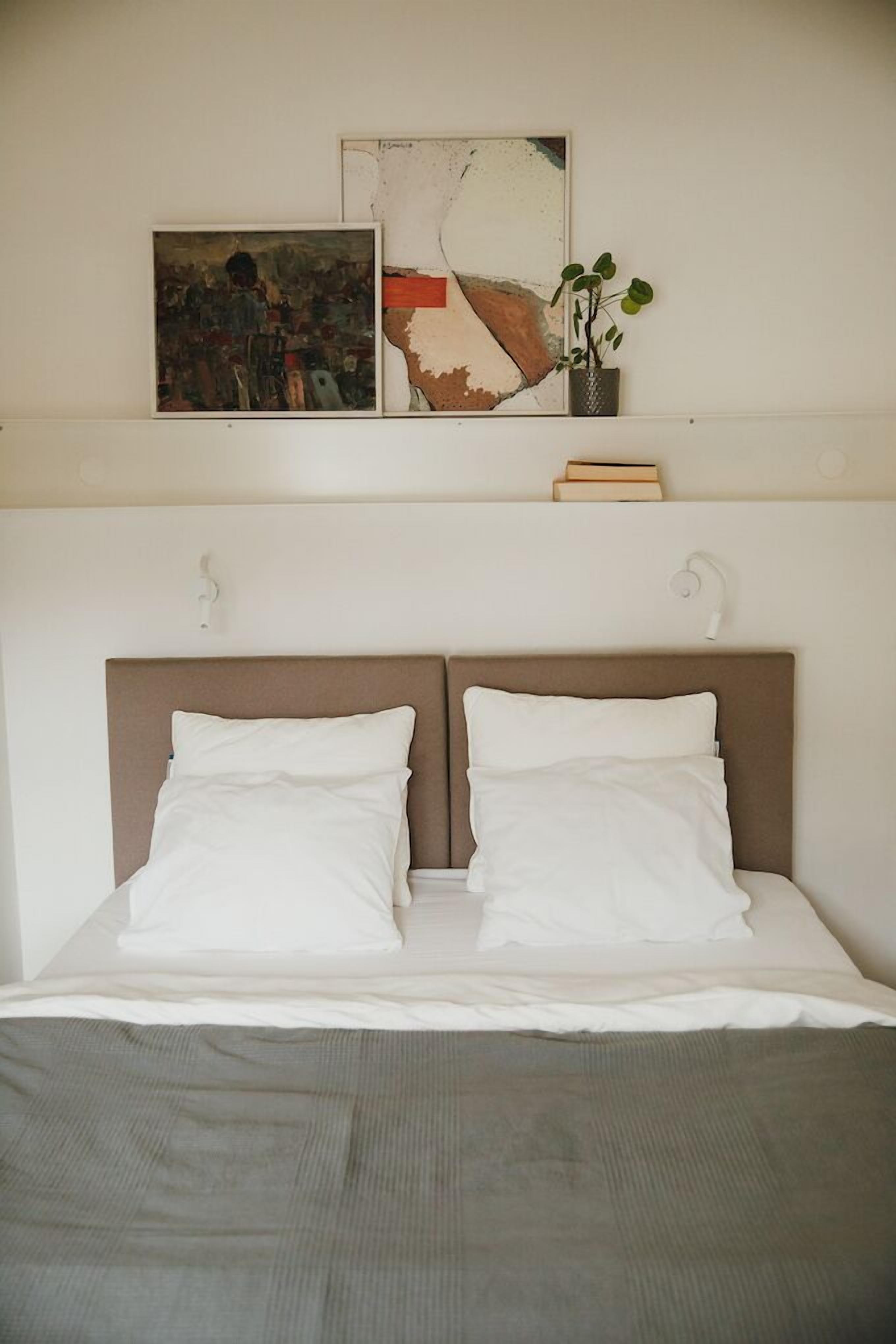Ein Doppelbett mit brauner Kopfteile, weißer Bettwäsche und einer grauen Tagesdecke. Darüber hängen zwei gerahmte Kunstwerke, und rechts daneben steht ein kleiner Topfpflanze.