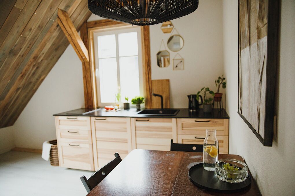 A kitchen with wooden cabinets, a black countertop, and a dining table with chairs. There is a window with natural light, a hanging light fixture, and a framed picture on the wall.