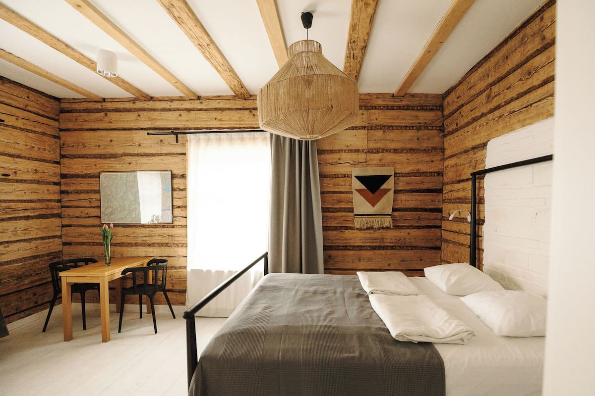 A bedroom with a wooden log wall, a bed with white linens, a desk with a chair, and a hanging light fixture.