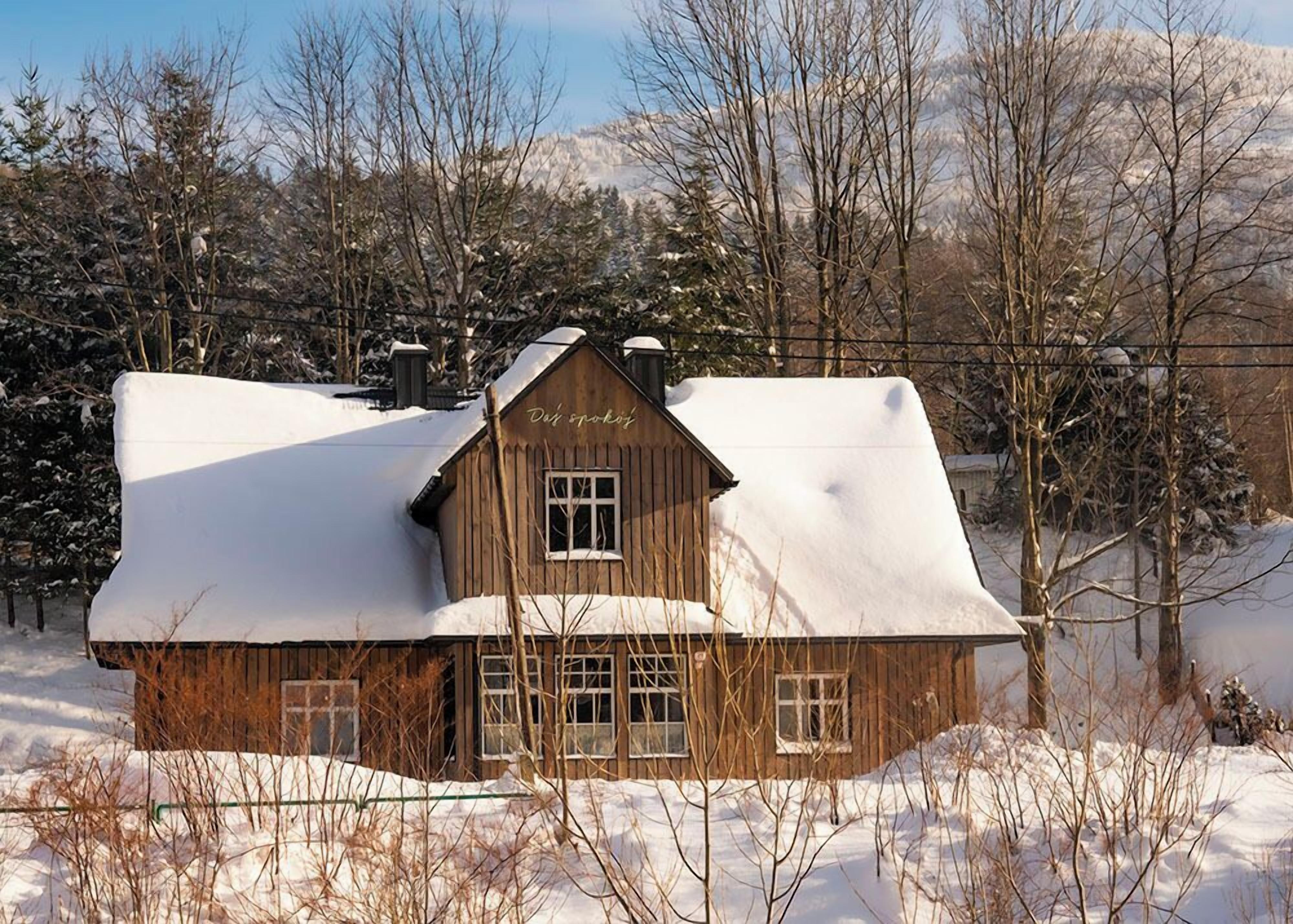 Une cabane en bois, dont le toit est recouvert de neige, entourée d’arbres et avec une chaîne de montagnes en arrière-plan.