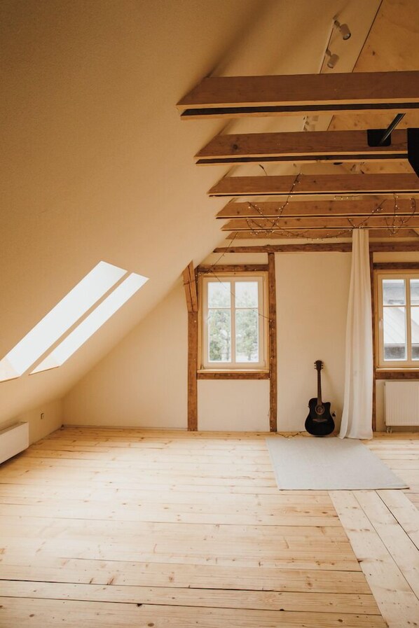 Una habitación con techo inclinado, vigas de madera, tragaluces, una ventana, una guitarra y un radiador.