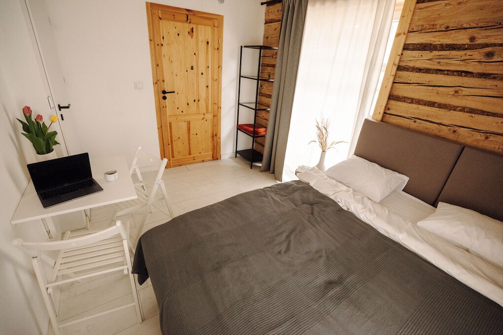 A bedroom with a wooden headboard, a bed with white linens, a desk with a laptop, a white chair, and a wooden wardrobe.