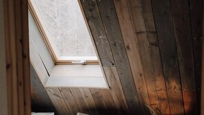 Een slaapkamer met een schuine kap, dakraam, bed met witte beddengoed, een klein houten krukje en een vaas met gedroogde planten.