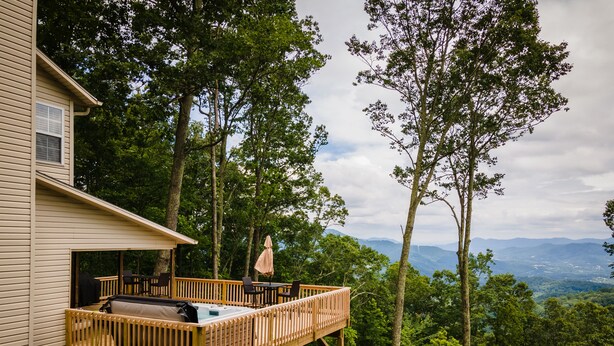 Una terraza de madera con mobiliario exterior, con vista a un paisaje boscoso y montañas distantes.