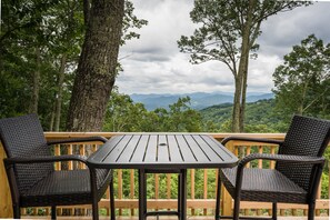 A deck with two wicker chairs and a table, overlooking a forested landscape with mountains in the distance.