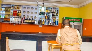 Zona de bar con estantes llenos de botellas de licor, una barra y una persona sentada en una mesa.