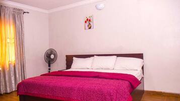 A bedroom with a wooden bed, white bedding, a red blanket, a wall-mounted fan, and a painting.