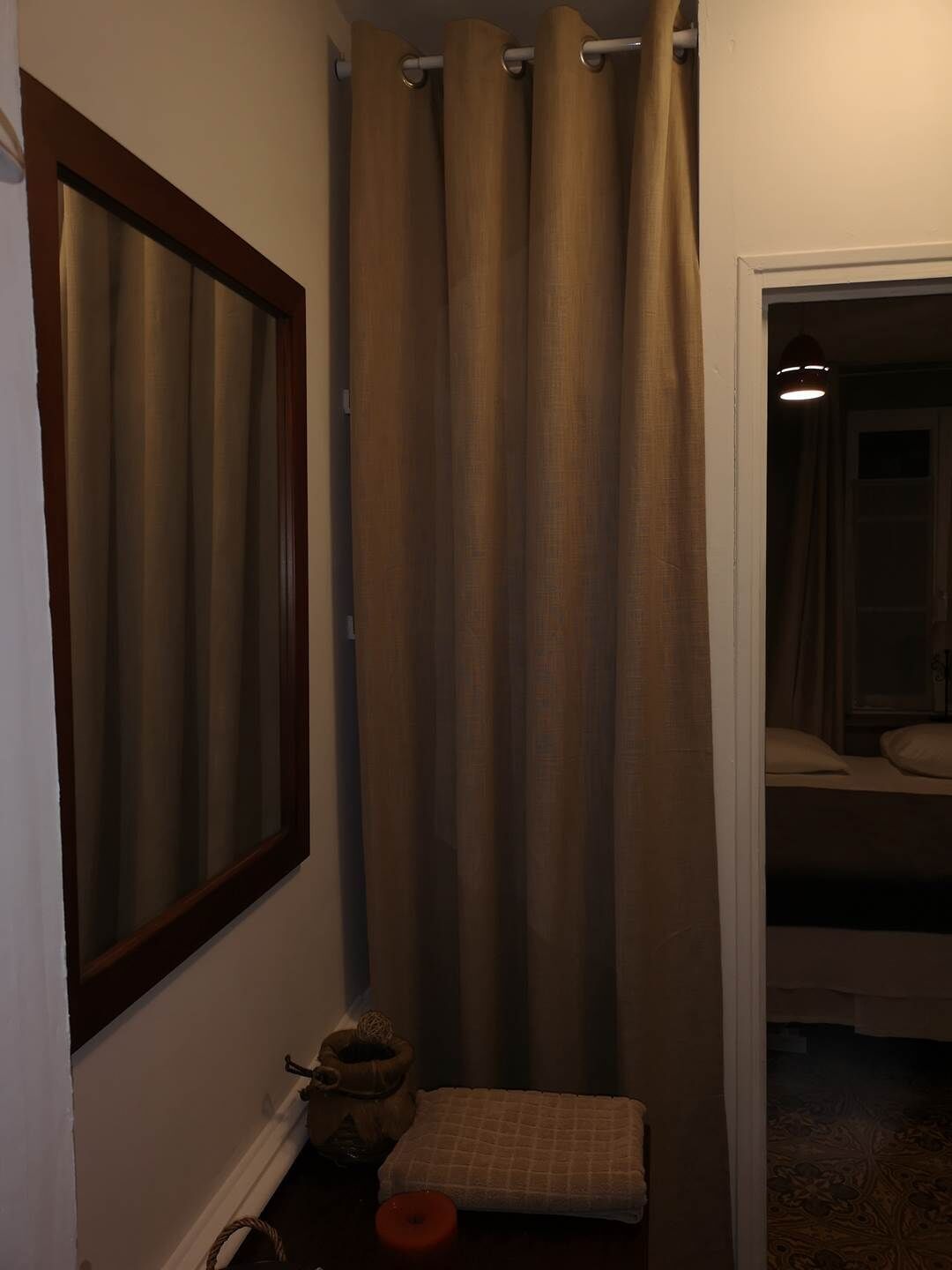 Een smalle gang met een spiegel met houten lijst, beige gordijnen, een klein vloerkleed en een nachtkastje met een lamp.