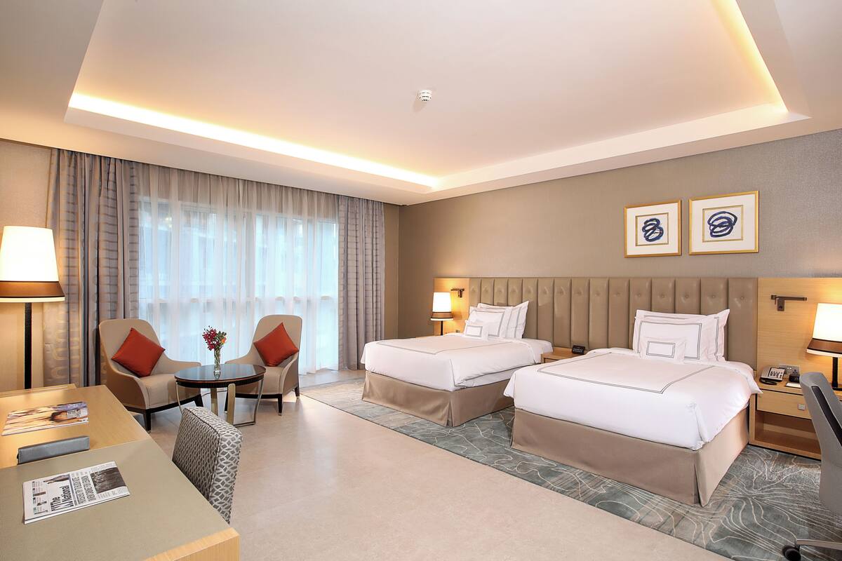 Une chambre d’hôtel avec deux lits, un coin salon avec une table et des chaises, et une grande fenêtre avec des rideaux.