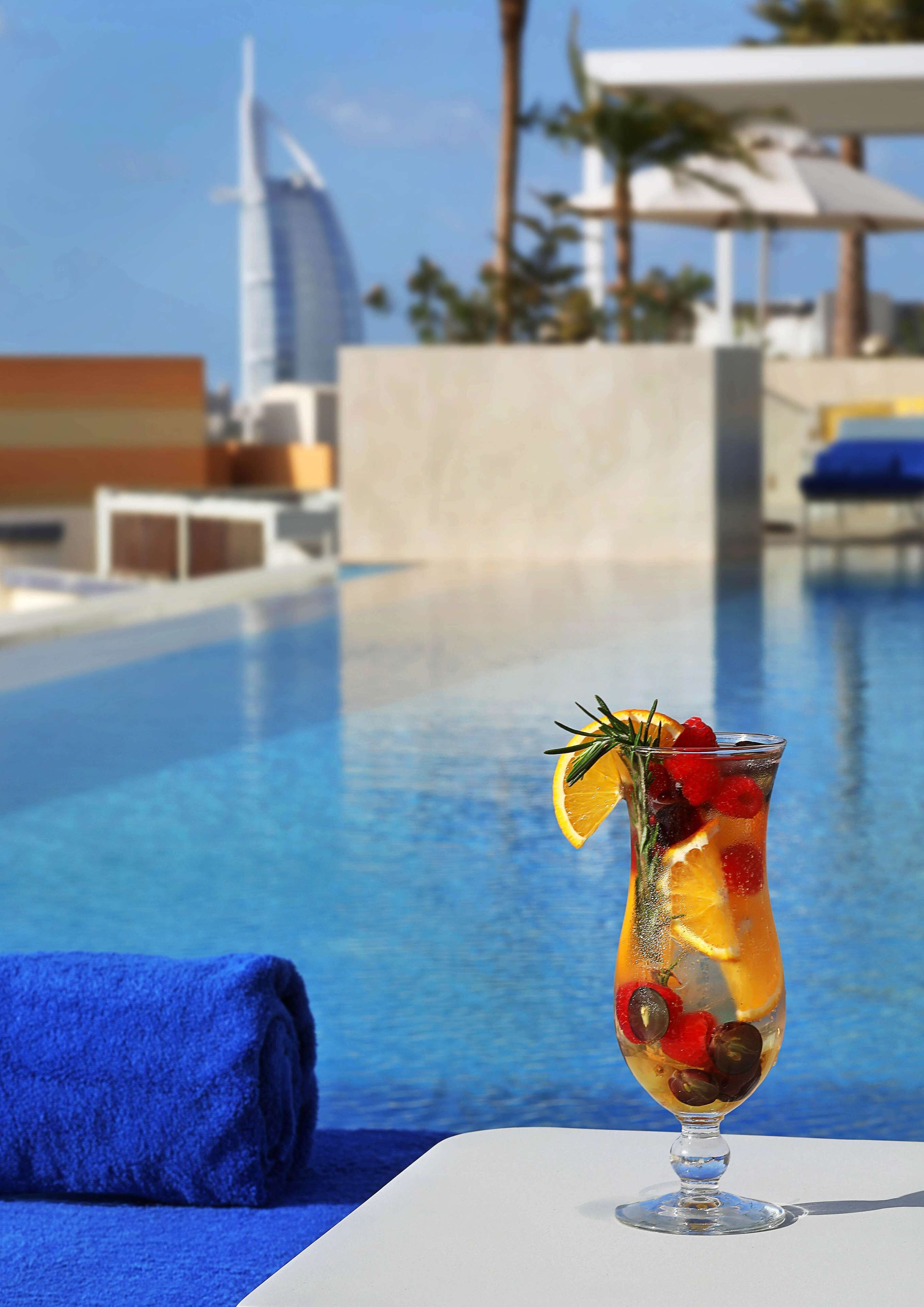 A clear view of a swimming pool with a glass of fruit cocktail on a table, a blue towel, and a modern building in the background.