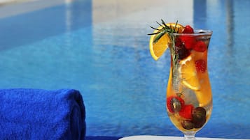 A clear view of a swimming pool with a glass of fruit cocktail on a table, a blue towel, and a modern building in the background.