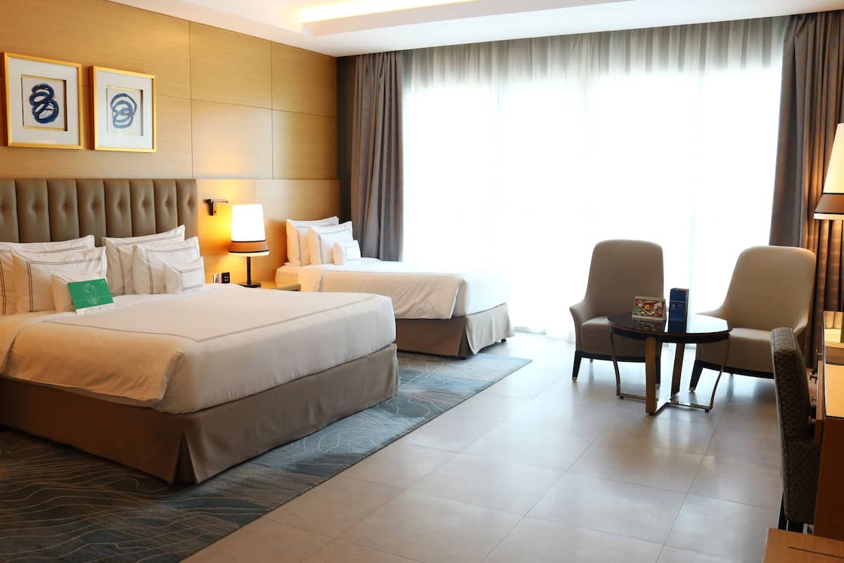 Une chambre d’hôtel avec deux lits, un coin salon avec une table et des chaises, et une grande fenêtre avec des rideaux.