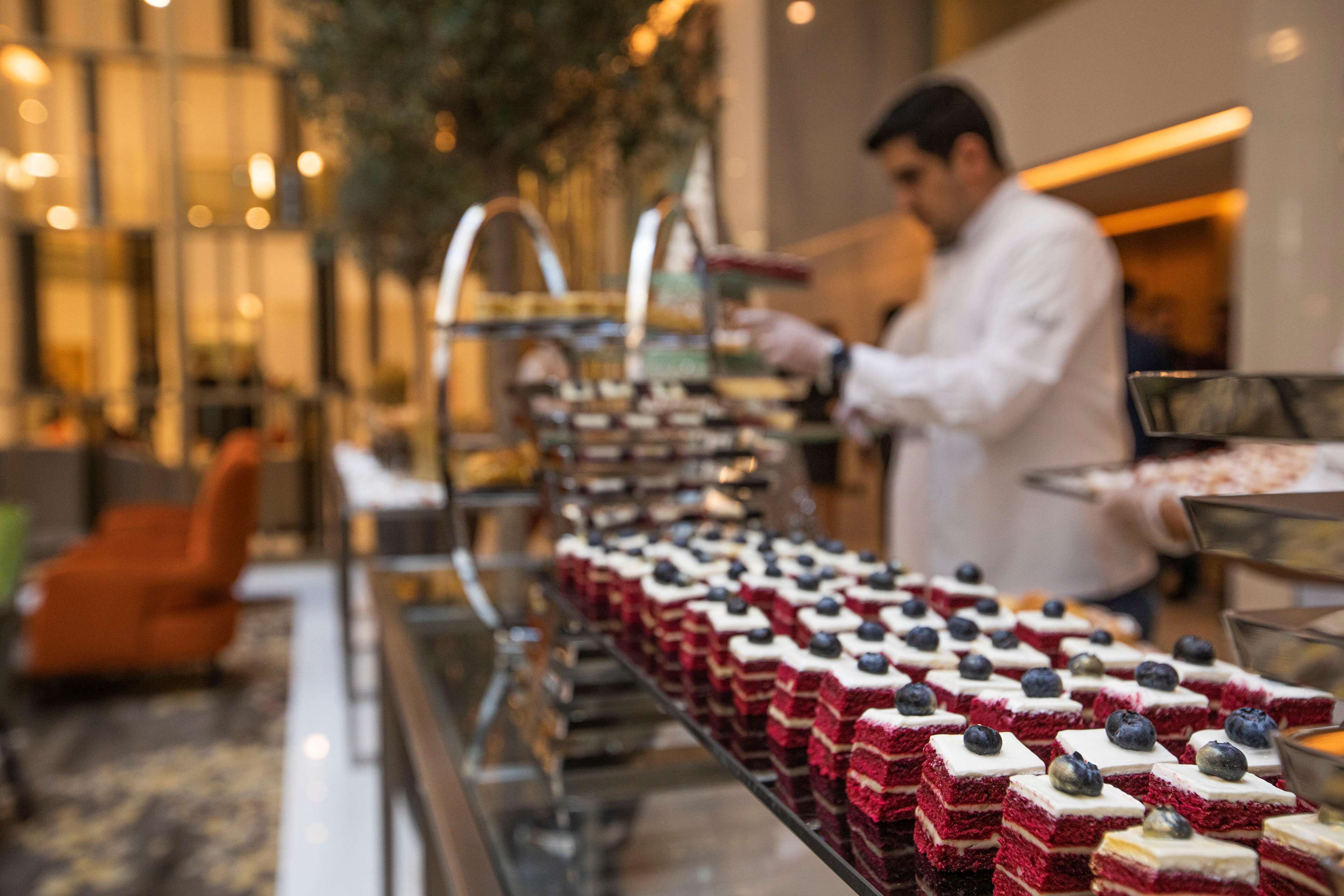 Buffet com sobremesas de veludo vermelho cobertas com mirtilos, uma pessoa a servir, e um interior moderno com guarda-corpo de vidro e cadeiras laranja.
