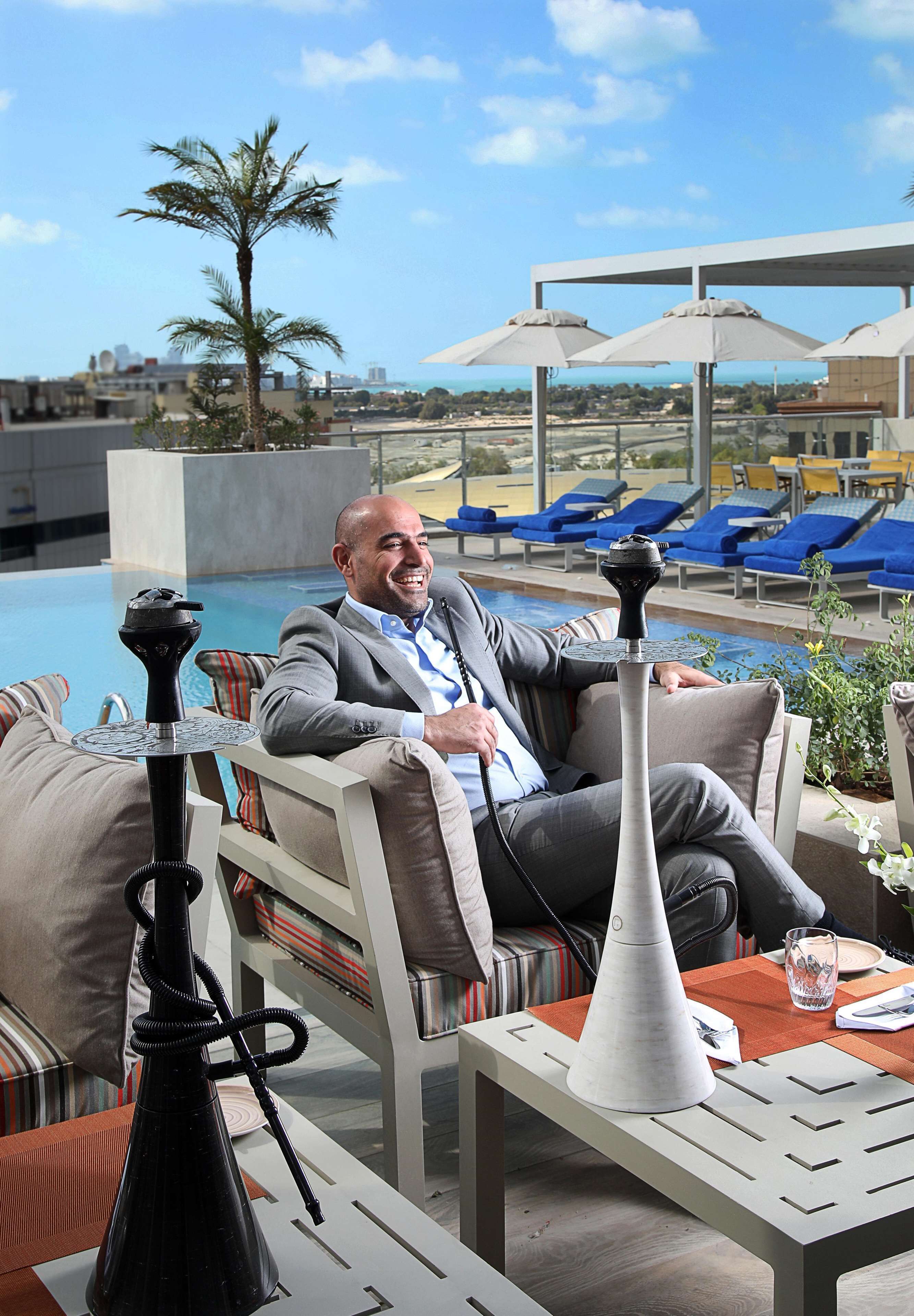 A man seated at a table with a hookah, overlooking a pool area with lounge chairs and umbrellas.