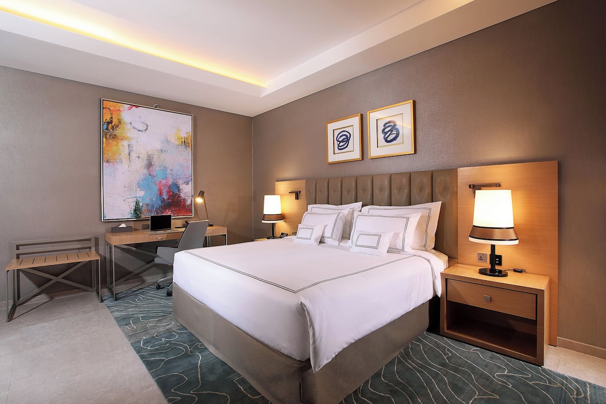Une chambre d’hôtel avec un grand lit, un bureau avec un ordinateur, une chaise, deux lampes de chevet et un grand tableau abstrait accroché au mur.