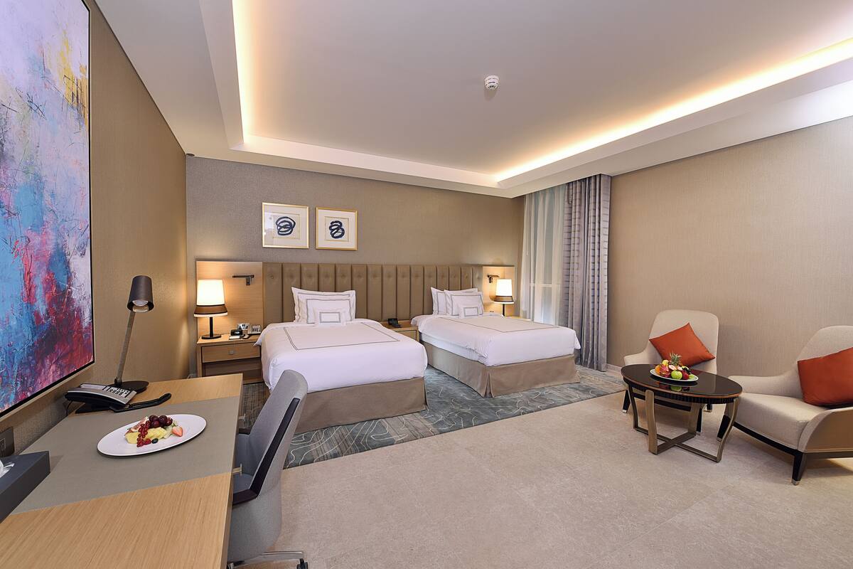 Une chambre d’hôtel avec deux lits, un bureau avec une assiette de fruits, une télévision et un coin salon avec une table et des chaises.