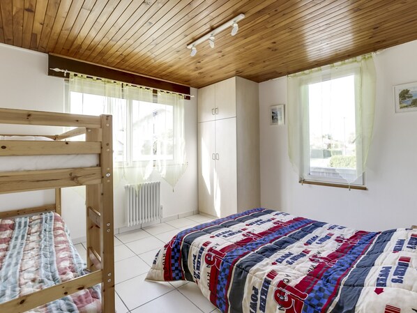 A bedroom with a bunk bed, a single bed with a patterned bedspread, a radiator, a window with curtains, and a wooden ceiling.
