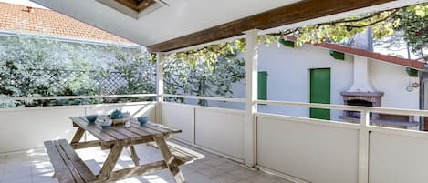 A covered patio with a wooden table and chairs, a tiled floor, and a view of a building with a green door and a chimney.