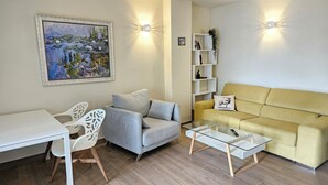 A modern living room with a sofa, a glass coffee table, a white desk, and a chair. There is a painting on the wall and a bookshelf with decorative items. - Apartment Lychee - Golden Star Javea close to Arenal beach. (Xàbia)