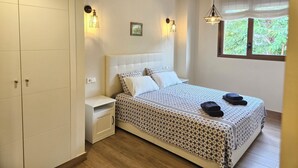 A bedroom with a bed, nightstand, wardrobe, wall-mounted lights, and a window with a view of greenery. - Apartment Lychee - Golden Star Javea close to Arenal beach. (Xàbia)