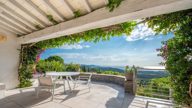 Une terrasse extérieure avec une table et des chaises blanches, entourée de verdure et d’un mur de pierre, offrant une vue sur un paysage pittoresque.