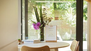 A wooden table with a vase of flowers and a welcome sign, set against a glass door with a view of greenery.