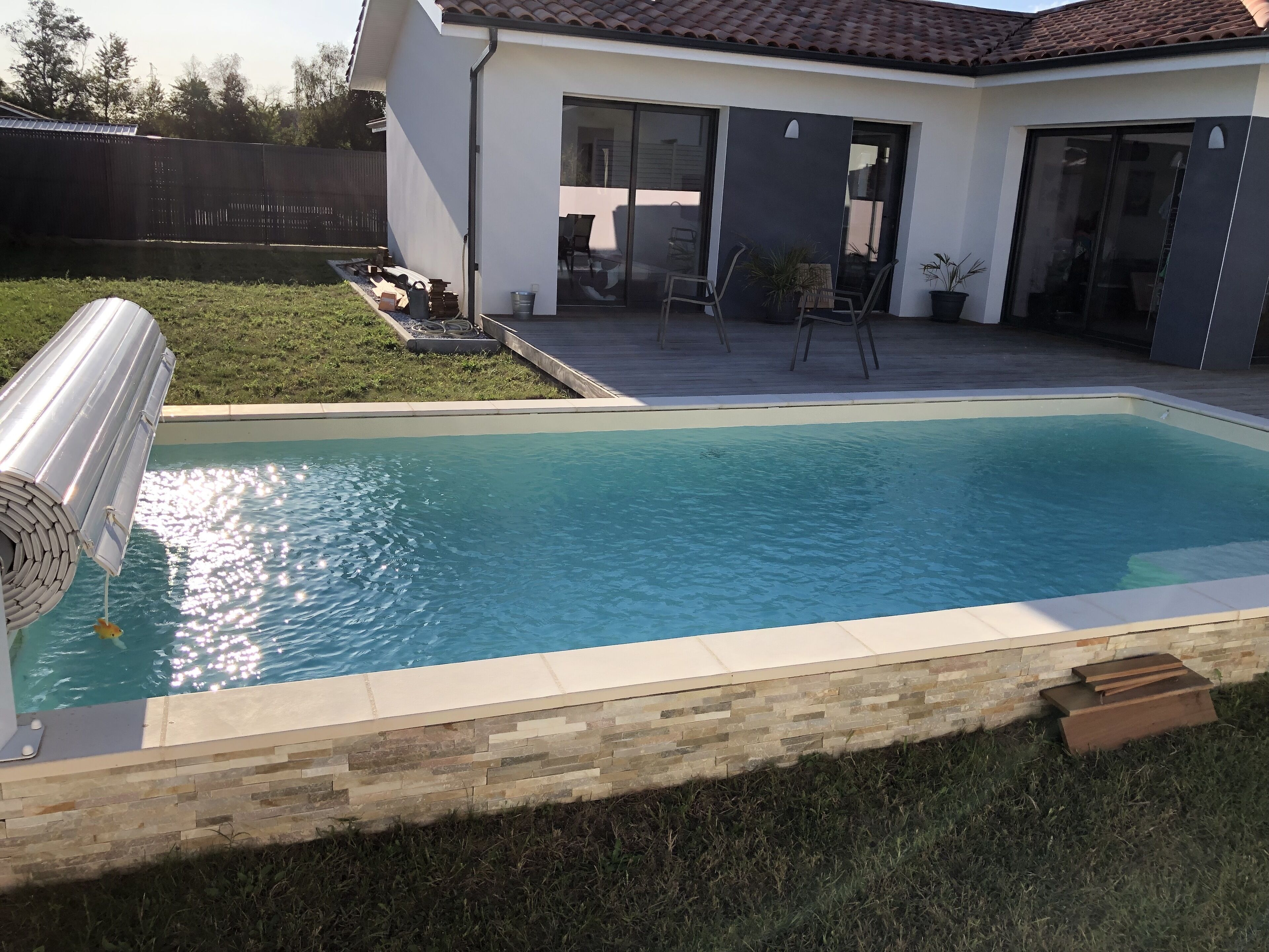 A rectangular swimming pool with a stone border, a patio area with chairs, and a modern house with a tiled roof in the background.