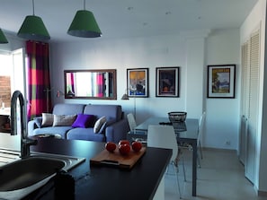 A modern living room with a sofa, a dining table with chairs, a kitchen area with a sink, and a window with curtains.