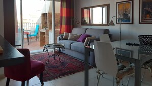 Una sala de estar con sofá, sillas, una mesa baja y una mesa de comedor. Hay un balcón con vistas a la ciudad.