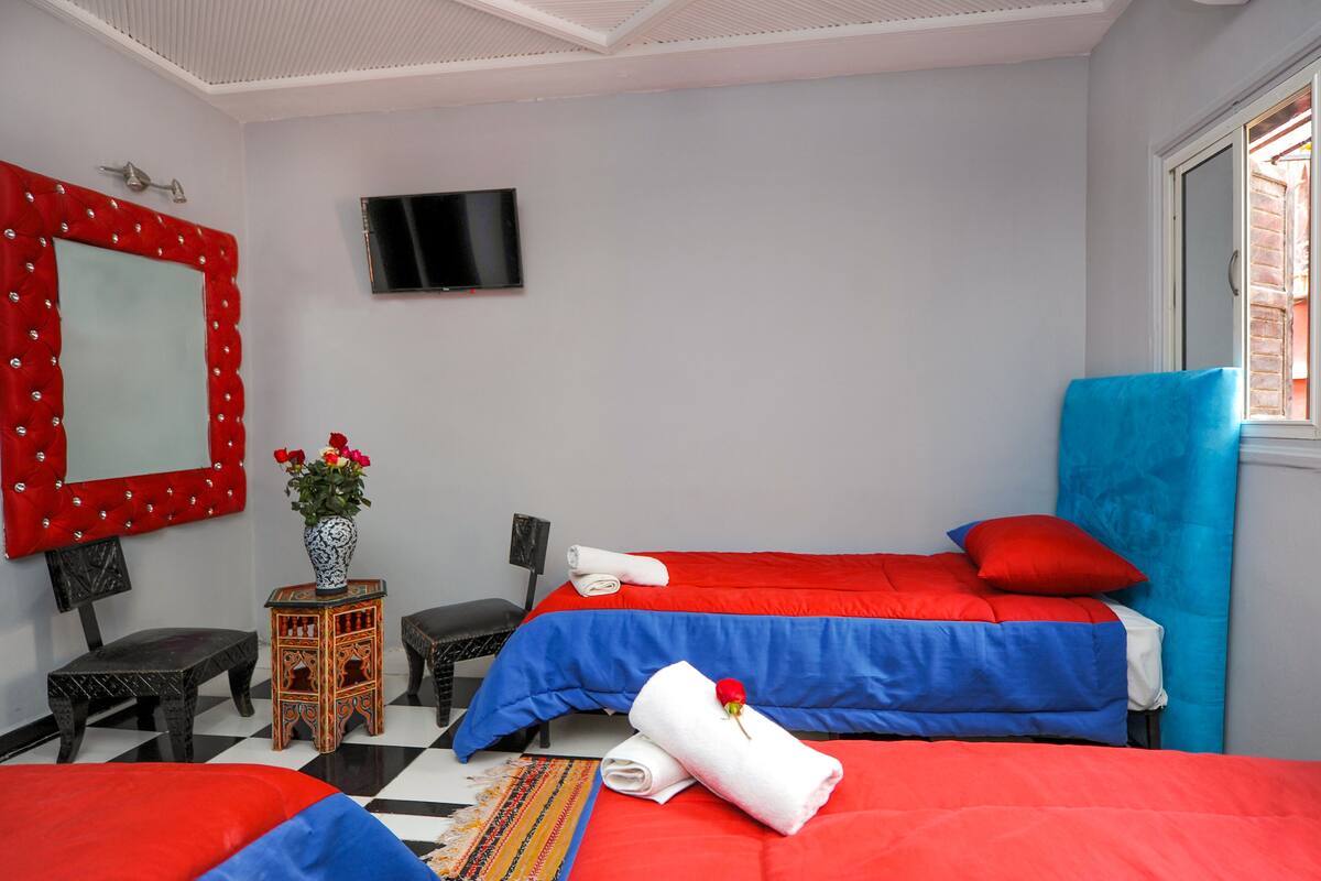 Habitación de hotel con dos camas, televisión, espejo, mesita con un florero y ventana con vistas.