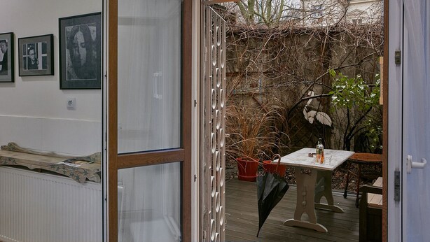 A room with a wooden floor, a radiator, a table with a bottle and glasses, and a door leading to an outdoor area with a wooden fence and plants.