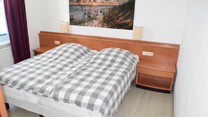 A neatly made bed with a checkered bedspread and pillows, a wooden headboard, a bedside table with a lamp, and a wall-mounted picture of a beach scene.