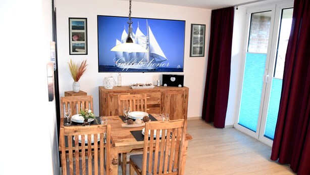 A dining area with a wooden table and chairs, a flat-screen TV mounted on the wall, and a view of the sea through a sliding glass door.