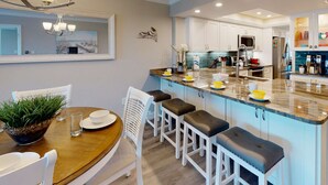 Une cuisine moderne avec un plan de travail en marbre, des armoires blanches et un coin repas comprenant une table en bois et des chaises blanches.