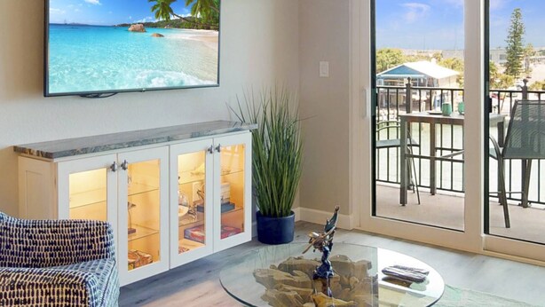 Un salon avec un téléviseur diffusant une scène de plage, une table basse en verre surmontée d’une sculpture, un meuble avec des portes en verre et une vue sur un balcon aménagé avec du mobilier d’extérieur.