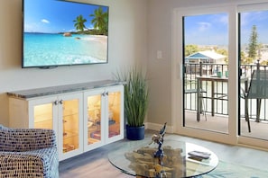 Una sala de estar con un televisor mostrando una escena de playa, una mesa de centro de vidrio con una escultura, un mueble bar con puertas de cristal y vistas a un balcón con mobiliario exterior.