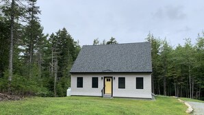 A single-story house with a grey roof and a yellow door, surrounded by a grassy yard and dense forest. - Newly built and fresh vacation home tucked in the woods of Mount Desert.  (Mount Desert)