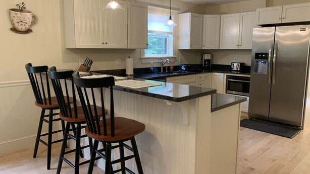 A modern kitchen with white cabinets, a stainless steel refrigerator, a bar area with stools, and pendant lighting. - Newly built and fresh vacation home tucked in the woods of Mount Desert.  (Mount Desert)