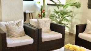 A seating area with two dark wicker sofas, each with a white cushion and a patterned pillow. A small table with a white cloth and yellow flowers is in the foreground.