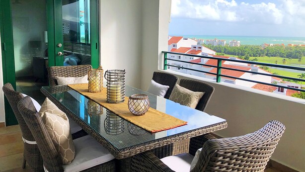 A balcony with wicker furniture and a glass table, overlooking a coastal view with buildings and greenery.