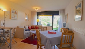 A cozy living room with a sofa, a dining table, and a TV. There are framed pictures on the walls and a window with curtains. - Lindenschlösschen & Schlossgarten (Garmisch-Partenkirchen)