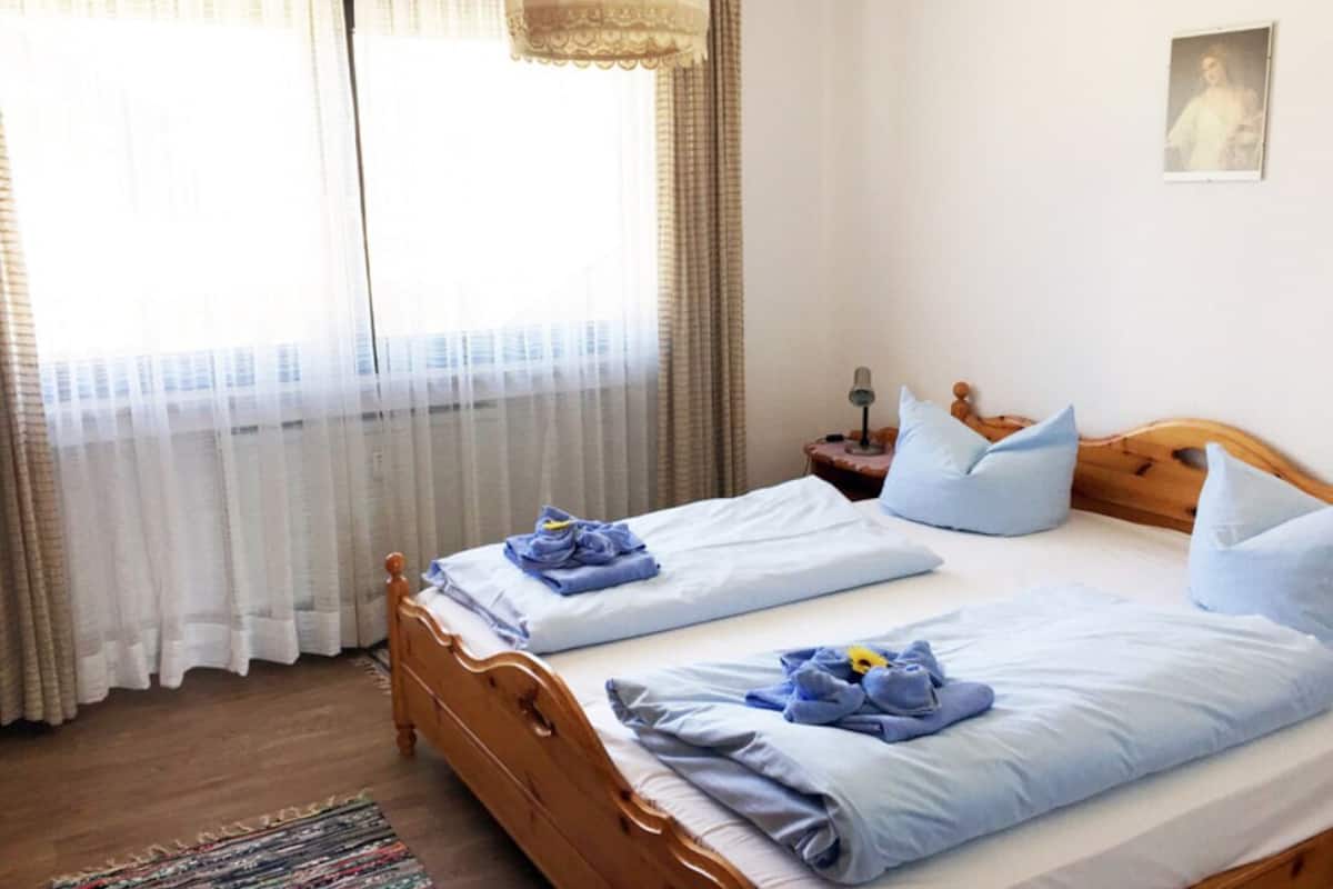 Uma cama de casal com roupa de cama azul, uma cabeceira de madeira, uma mesa de cabeceira com abajur e uma janela com cortinas leves.