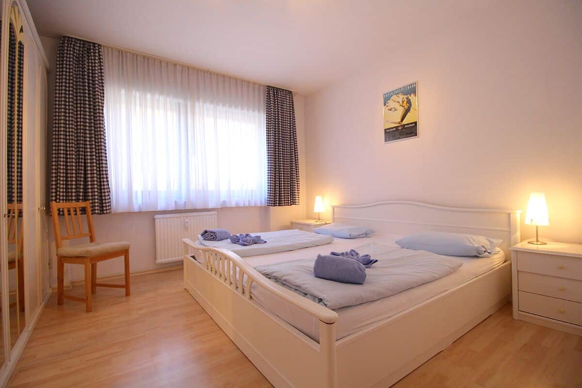 Quarto com uma cama grande, duas mesas de cabeceira com abajures, uma cadeira de madeira e uma janela com cortinas.