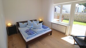 Un dormitorio con una cama de madera, dos mesitas de noche, una silla y un amplio ventanal con vistas a un jardín y edificios.